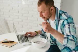 hermoso sonriente hombre en camisa sentado en cocina a hogar a mesa trabajando en línea en ordenador portátil foto
