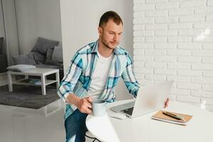 handsome smiling man in shirt sitting in kitchen at home at table working online on laptop photo