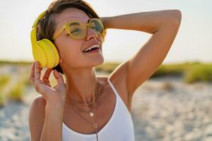 happy smiling woman listening to music in colorful yellow headphones on sunny beach in summer photo