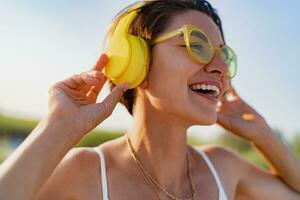 happy smiling woman listening to music in colorful yellow headphones on sunny beach in summer photo