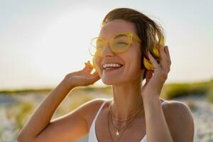 happy smiling woman listening to music in colorful yellow headphones on sunny beach in summer photo