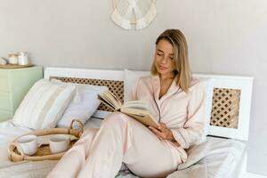 pretty smiling woman relaxing at home on bed in morning in pajamas reading book photo