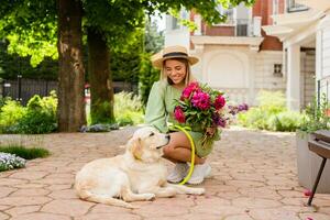 beautiful young woman in summer style outfit smiling happy walking with flowers and dog photo