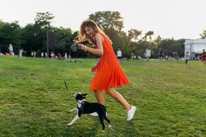 joven contento sonriente mujer en naranja vestir teniendo divertido jugando con perro en parque foto