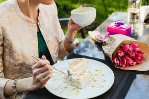 young stylish woman sitting in cafe, holding drinking cup cappuccino, eating tasty cake photo