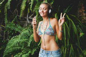 joven sexy mujer en bikini parte superior y mezclilla bermudas, escuchando a música en auriculares foto