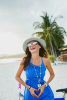 joven atractivo sonriente mujer en azul vestir caminando en tropical playa con bicicleta foto