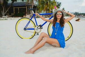 joven atractivo sonriente mujer en azul vestir caminando en tropical playa con bicicleta foto