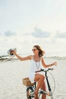 young attractive smiling woman in white dress riding on tropical beach on bicycle photo