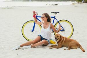 young beautiful woman sitting on sand on beach, holding vintage bicycle photo