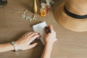 young stylish beautiful woman sitting at table in resort hotel room, writing a letter, holding pen photo