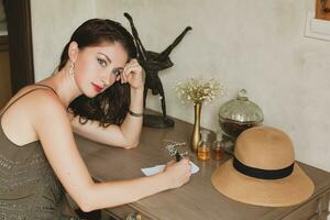 young stylish beautiful woman sitting at table in resort hotel room, writing a letter, holding pen photo
