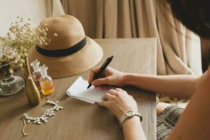 young stylish beautiful woman sitting at table in resort hotel room, writing a letter, holding pen photo