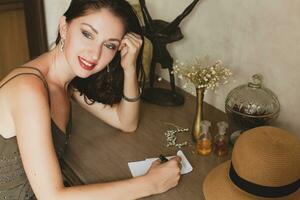 young stylish beautiful woman sitting at table in resort hotel room, writing a letter, holding pen photo