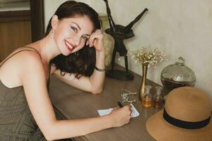 young stylish beautiful woman sitting at table in resort hotel room, writing a letter, holding pen photo