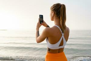 pretty young smiling woman doing sports in the morning in stylish sport outfit sportswear, using smartphone photo