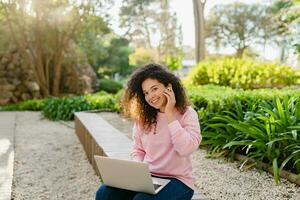 pretty curly young woman sitting in park working on laptop remote job photo