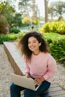 pretty curly young woman sitting in park working on laptop remote job photo