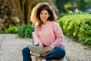 pretty curly young woman sitting in park working on laptop remote job photo