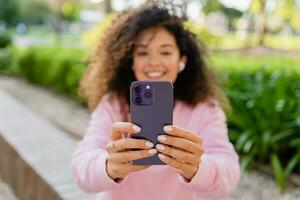 bonito hermosa Rizado joven mujer sentado en parque utilizando smatphone foto