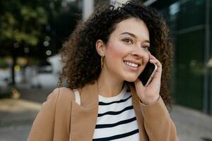 bonito Rizado sonriente mujer caminando en ciudad calle en elegante chaqueta, hablando en teléfono inteligente foto