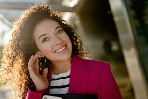 pretty curly woman in city street in stylish pink jacket, student education photo