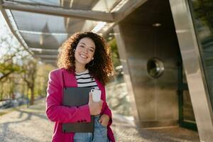 pretty curly woman in city street in stylish pink jacket, student education photo