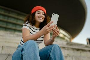 pretty curly smiling woman sitting in city street in striped t-shirt and knitted red hat, using smartphone photo