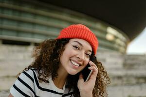 pretty curly smiling woman sitting in city street in striped t-shirt and knitted red hat, using smartphone photo