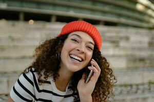 pretty curly smiling woman sitting in city street in striped t-shirt and knitted red hat, using smartphone photo