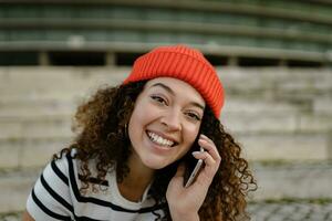 bonito Rizado sonriente mujer sentado en ciudad calle en a rayas camiseta y de punto rojo sombrero, utilizando teléfono inteligente foto