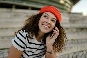 bonito Rizado sonriente mujer sentado en ciudad calle en a rayas camiseta y de punto rojo sombrero, utilizando teléfono inteligente foto