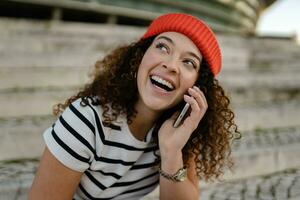 bonito Rizado sonriente mujer sentado en ciudad calle en a rayas camiseta y de punto rojo sombrero, utilizando teléfono inteligente foto