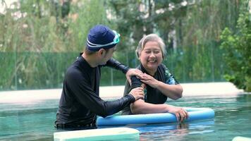 jong trainer helpen senior vrouw in aqua aerobics en werken uit in de zwembad. oud vrouw en volwassen Mens aan het doen aqua aerobics oefening in zwemmen zwembad, ouderen sport, en actief levensstijl concept. video