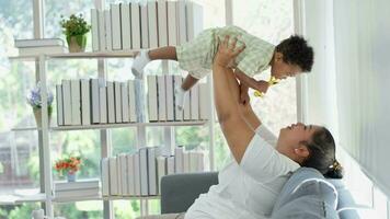 Happy Asian mother plays with little infant toddler child on living room sofa. Parent and little kid relaxing at home. Parents and toddler child girl enjoying spending time at home. Childcare concept video