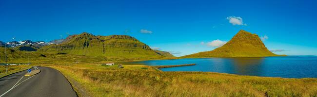 Panoramic over wonderful waterfall named Kirkjufellsfoss with a Kirkjufell church like iconic mount in Western Iceland, paved ring road at blue sky and sunny day photo