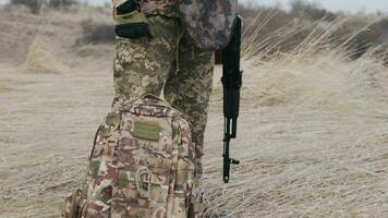 Professional soldier with weapon in military uniform stands on guard with autumn field and grey sky in background. Troop on the roadblock. Combatant in full ammunition. Concept of war video