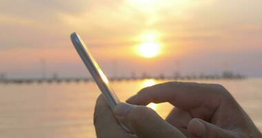 homme dactylographie sur téléphone intelligent par mer à le coucher du soleil video