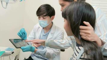 asiatisk manlig pediatrisk tandläkare förklarar tänder röntgen skanna till flicka och henne mor för tandvård hygien från läsplatta i dental klinik, professionell ortodontisk oral läkare i en unge sjukvård sjukhus. video