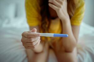 Young asian young woman feeling shock after look at positive Pregnancy test. Disappointed unplanned pregnancy girl sit on bed with upset and sad face for problem don't want to pregnant. photo