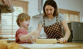 padre y madre enseñando bebé hijo amasadura masa en cocina mostrador a hogar. padres y chico niño disfrutar y divertido adentro actividad Cocinando juntos. foto