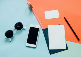 Creative flat lay style workspace desk with smartphone, blank envelope, sunglasses and masking tape on modern colorful background photo