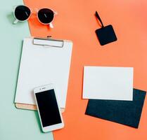 Creative flat lay of workspace desk with smartphone, clipboard, envelope, tag and sunglasses on minimal color background photo