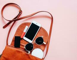 Flat lay of brown leather woman bag open out with cosmetics, accessories and smartphone on pink background photo