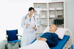 Asian doctor in white suit take notes while discussing and Asian elderly, man patient who lying on bed with receiving saline solution in hospital photo