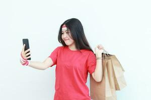 retrato de hermosa asiático mujer vistiendo rojo atuendo celebrando Indonesia independencia día por gesticulando que lleva un montón de compras pantalones mientras mirando a célula teléfono con sonriente expresión foto