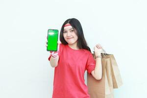 portrait of beautiful asian woman wearing red outfit celebrating Indonesia independence day by gesturing carrying lots of shopping bags and cell phone with green screen photo