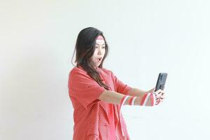 retrato de hermosa asiático mujer vistiendo rojo atuendo celebrando Indonesia independencia día mirando a móvil teléfono con sorprendido expresión foto