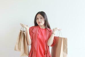 retrato de hermosa asiático mujer vistiendo rojo atuendo gesticulando que lleva un montón de compras pantalones con pensando expresión foto