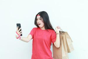 portrait of beautiful asian woman wearing red outfit celebrating Indonesia independence day by gesturing carrying lots of shopping bags while looking at mobile phone with surprised expression photo